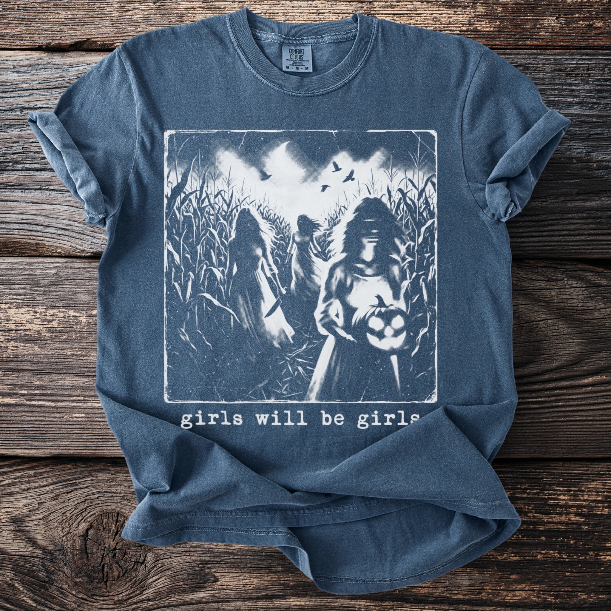 Blue t-shirt with spooky graphic of figures in a cornfield and text "girls will be girls."