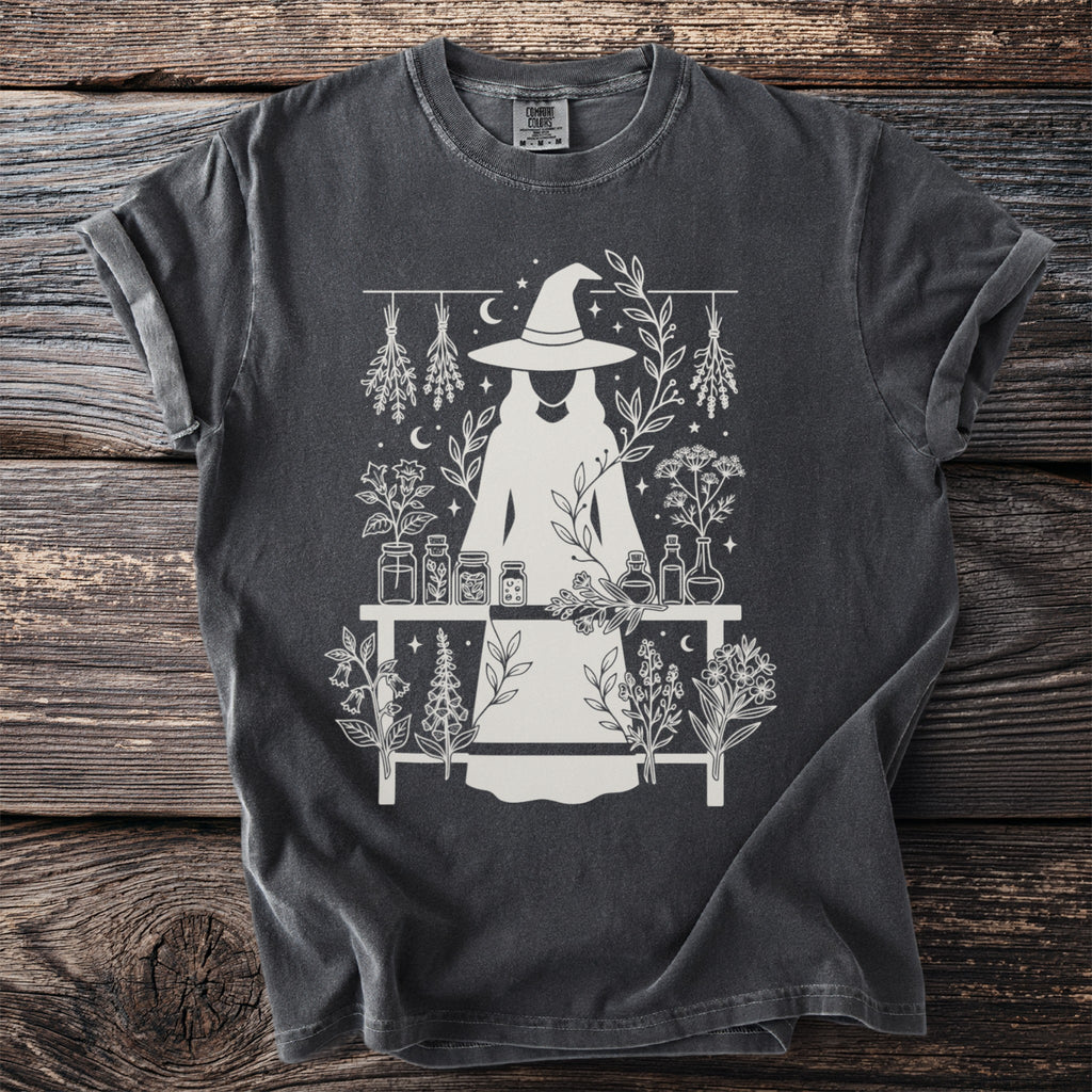 Dark gray shirt with white graphic of a witch and botanical elements.