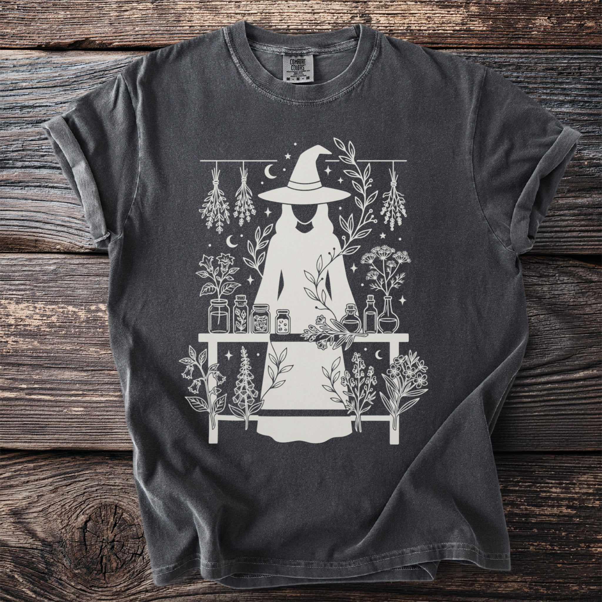 Dark gray shirt with white graphic of a witch and botanical elements.
