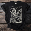 Black t-shirt with a cream folk-cut print: Krampus hangs one glass ornament on a snowy fir, chains and bells at his waist.