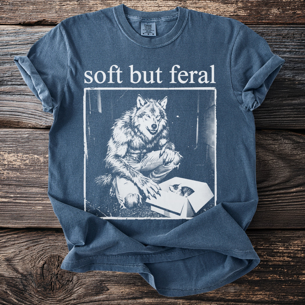 Blue t-shirt with "soft but feral" text and a werewolf looking at a kitten in a box.