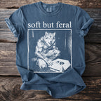 Blue t-shirt with "soft but feral" text and a werewolf looking at a kitten in a box.