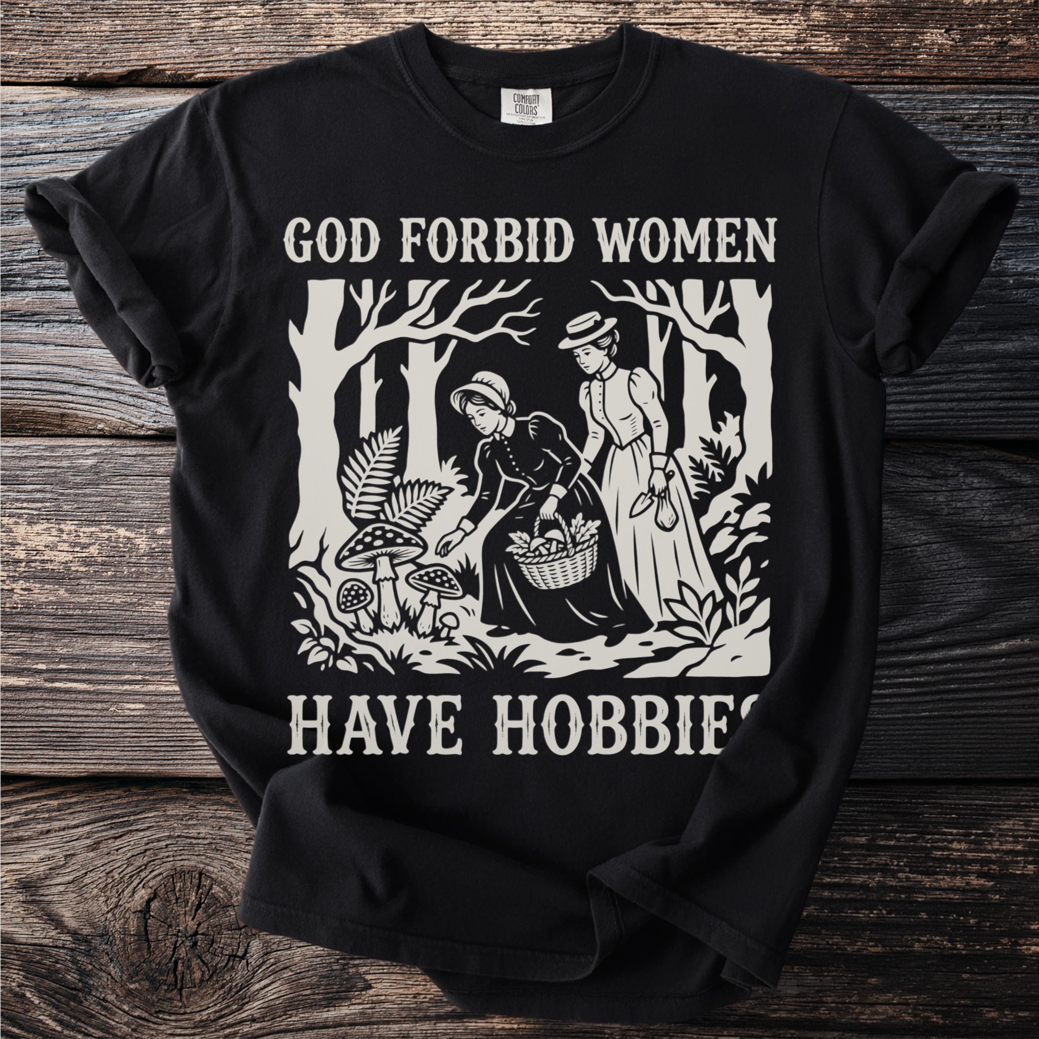 Black shirt with "God Forbid Women Have Hobbies" text and forest scene illustration.