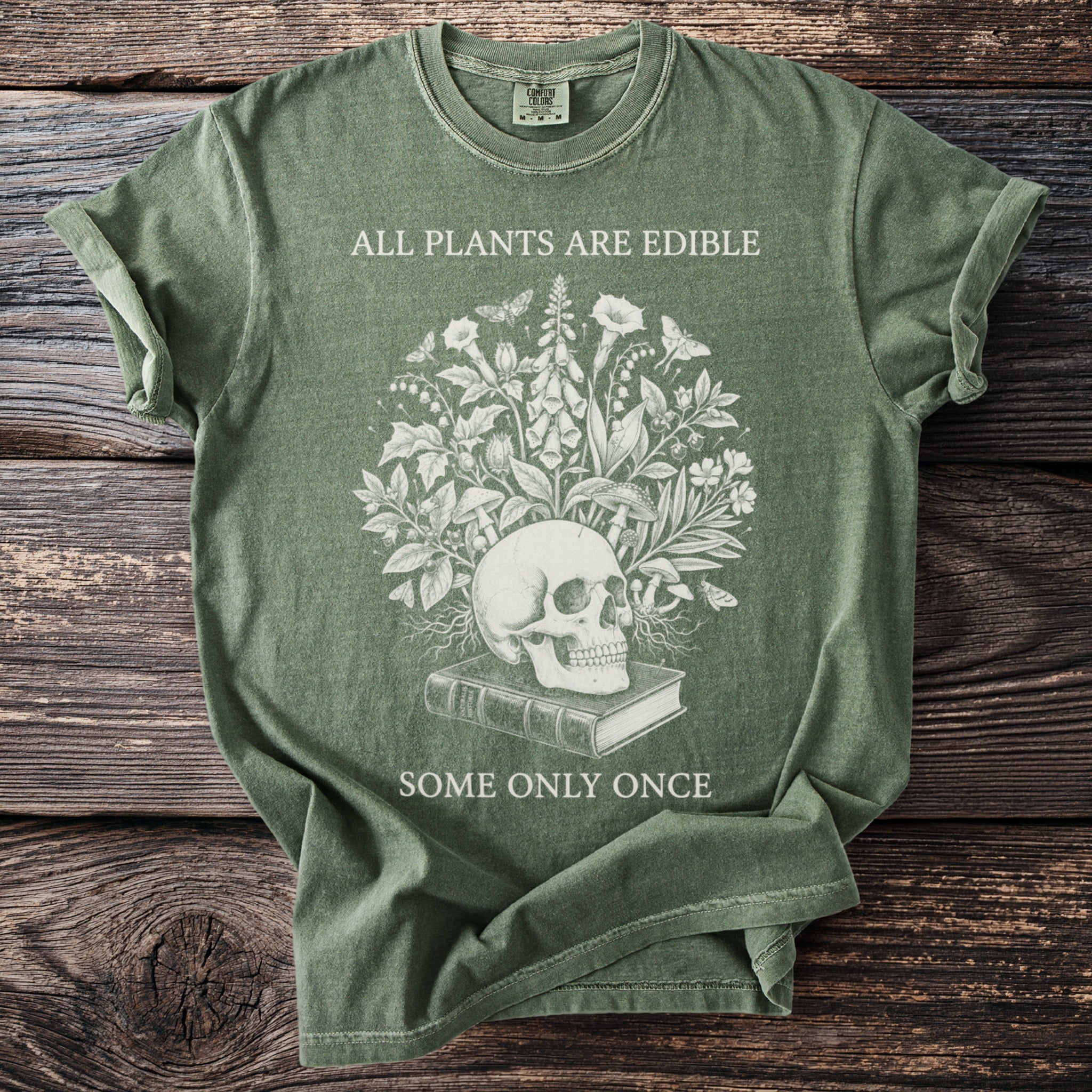 Green t-shirt with skull, plants, mushrooms, book, and text "ALL PLANTS ARE EDIBLE SOME ONLY ONCE".