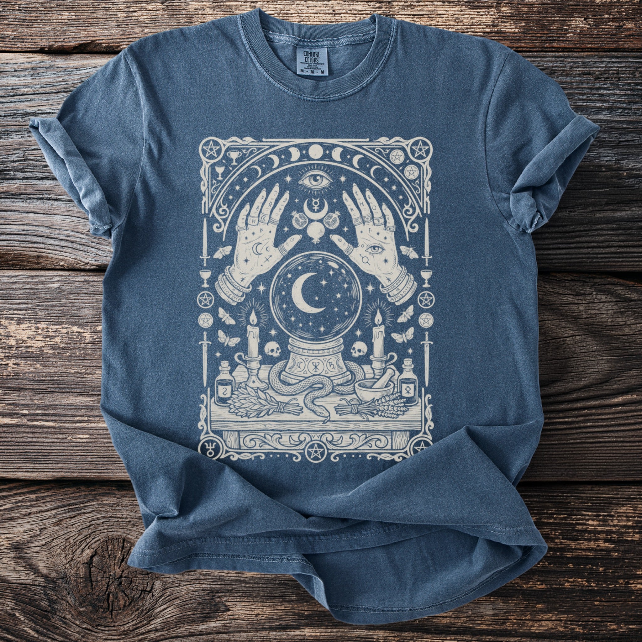 Blue t-shirt with mystical illustration of hands, crystal ball, moon, candles and occult symbols.