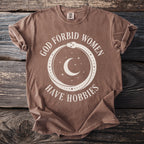 Brown t-shirt with text "GOD FORBID WOMEN HAVE HOBBIES" and celestial imagery.