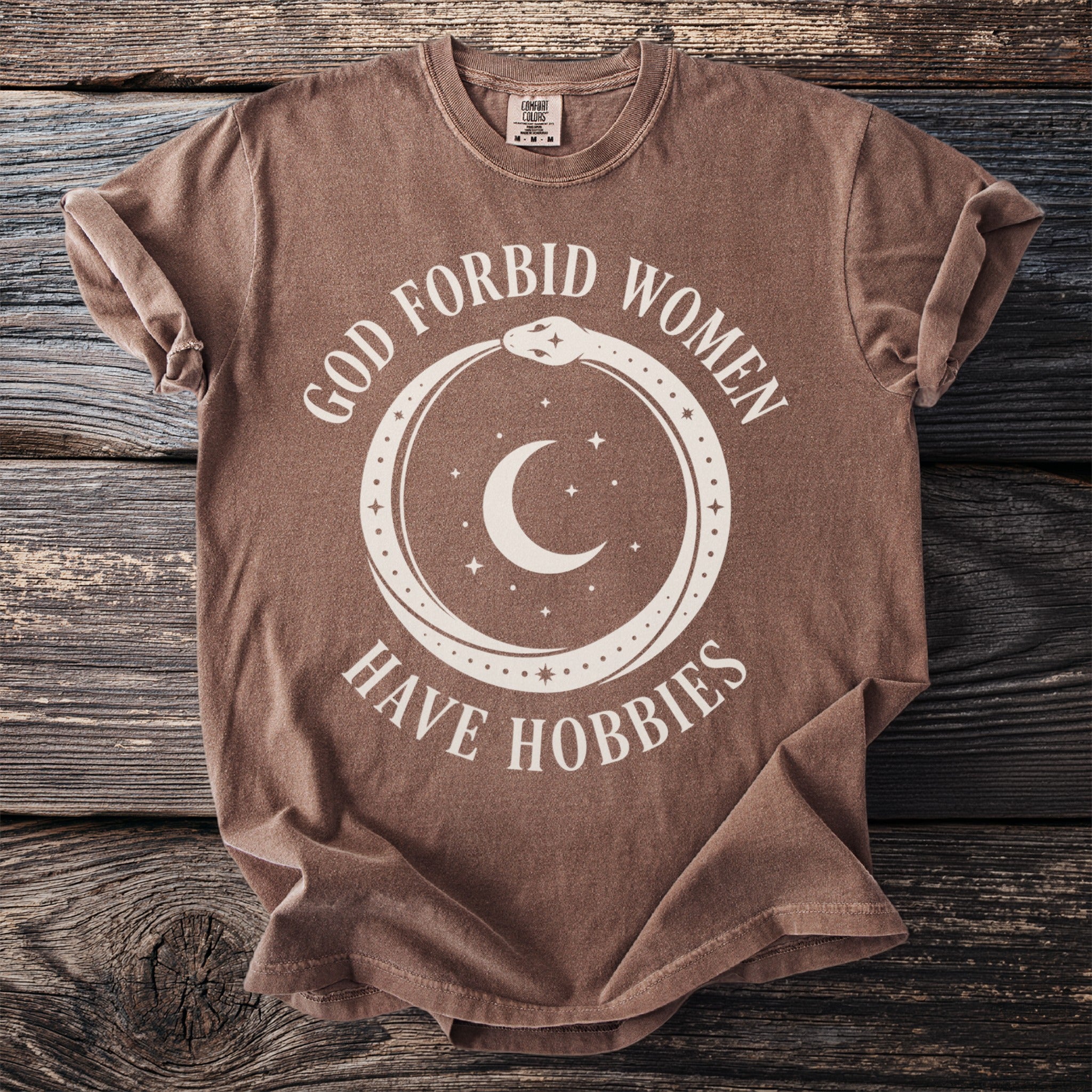 Brown t-shirt with text "GOD FORBID WOMEN HAVE HOBBIES" and celestial imagery.