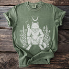 Green t-shirt with a white illustration of a werewolf sitting among plants and mushrooms.