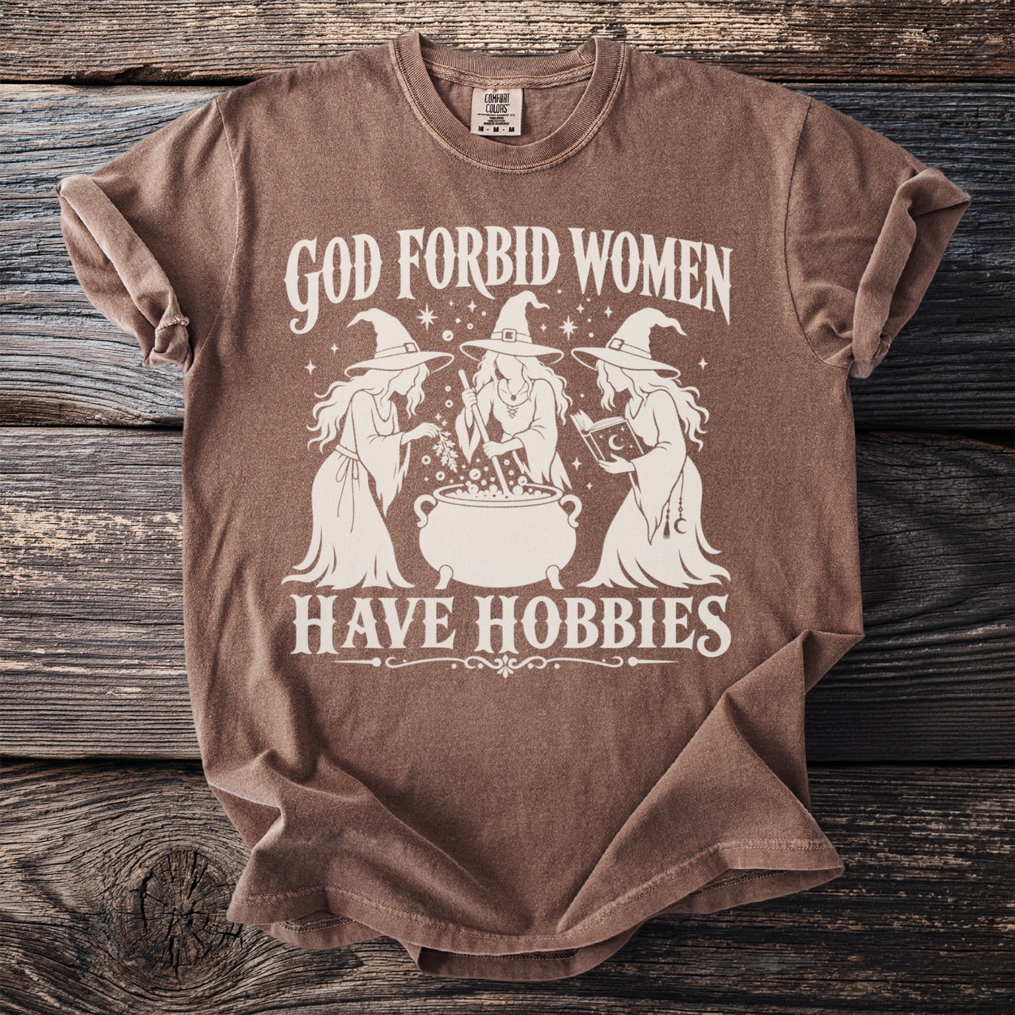 Brown t-shirt with white graphic text "God Forbid Women Have Hobbies" and three witches.