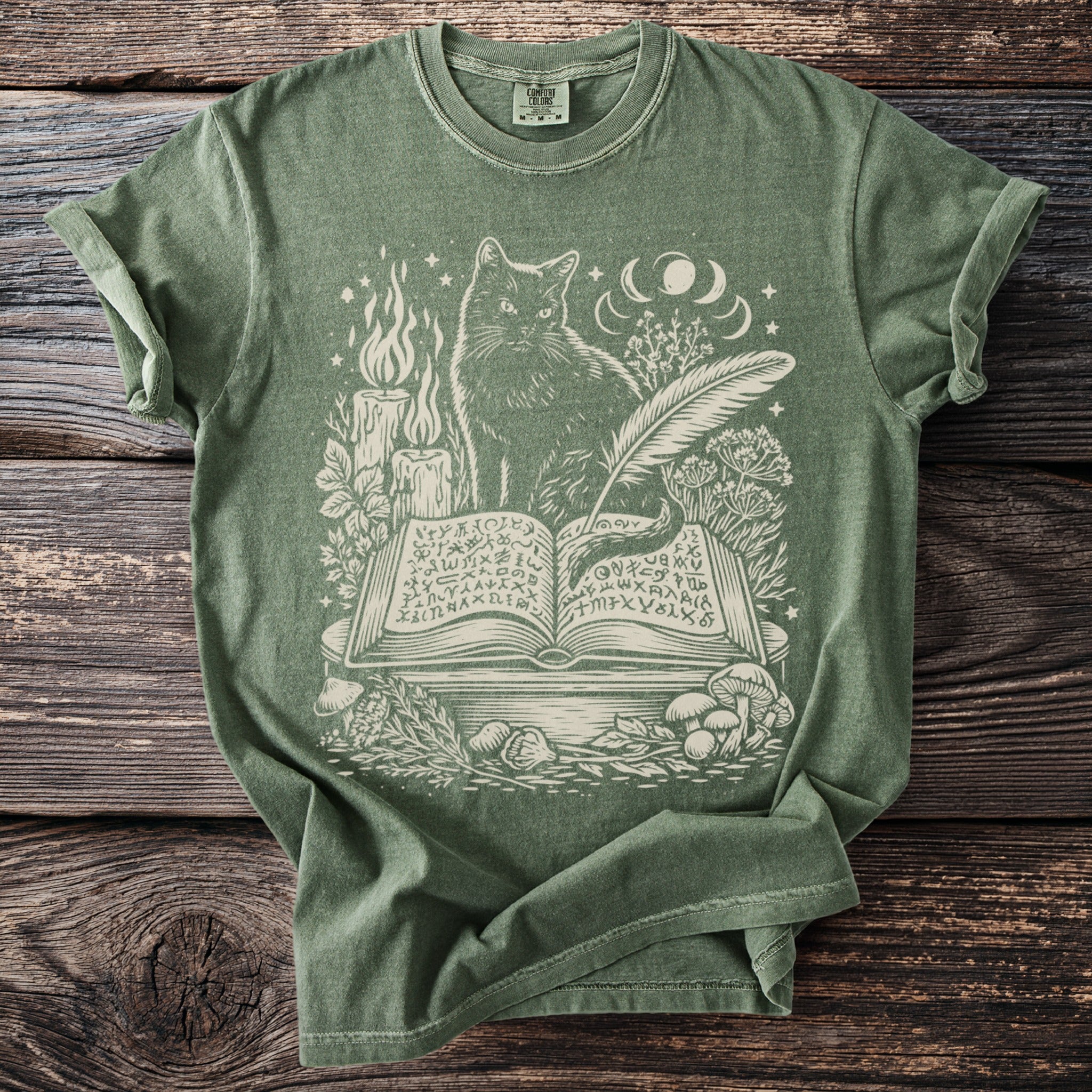Green t-shirt with cream print of cat on spellbook, candles, and mushrooms.