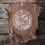 Brown t-shirt with white mystical book stack, moth, key, candle, and moon graphic.