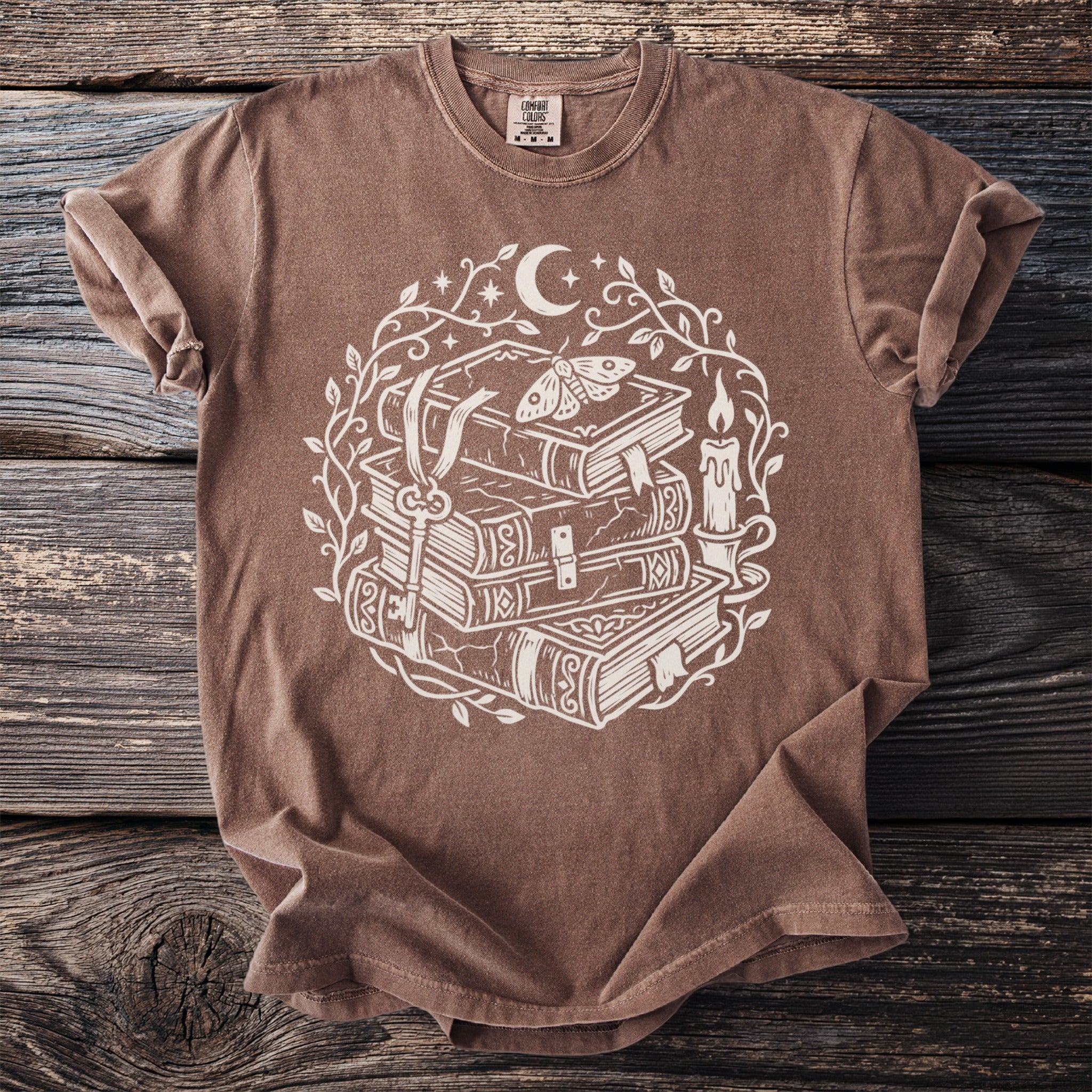 Brown t-shirt with white mystical book stack, moth, key, candle, and moon graphic.