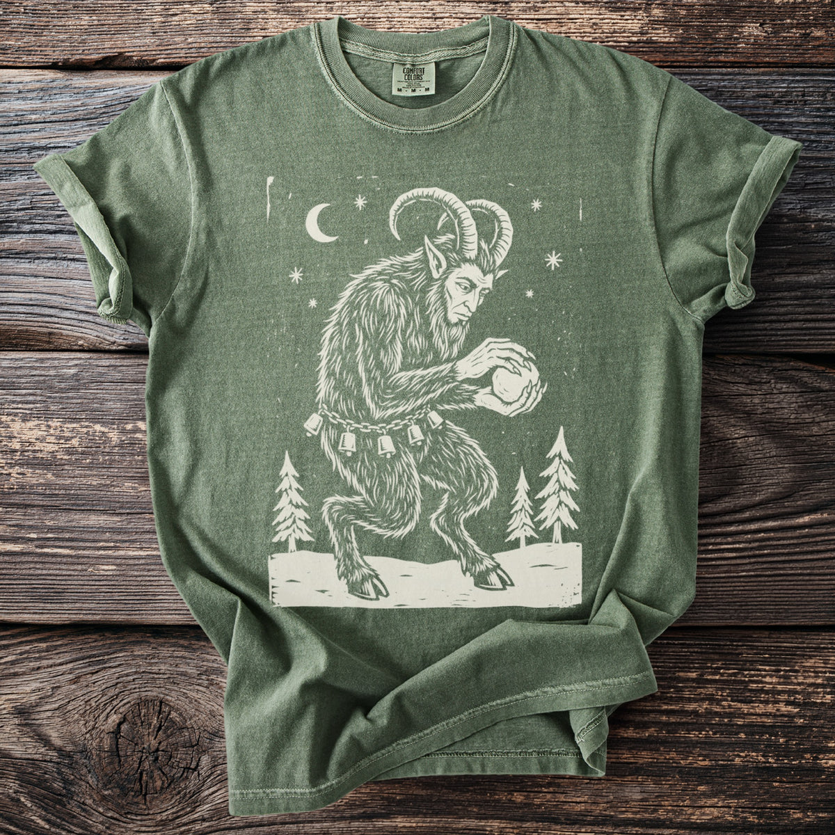 Moss green t-shirt with a cream folk-cut print: Krampus crouches among snowy pines, packing a snowball between his clawed hands.