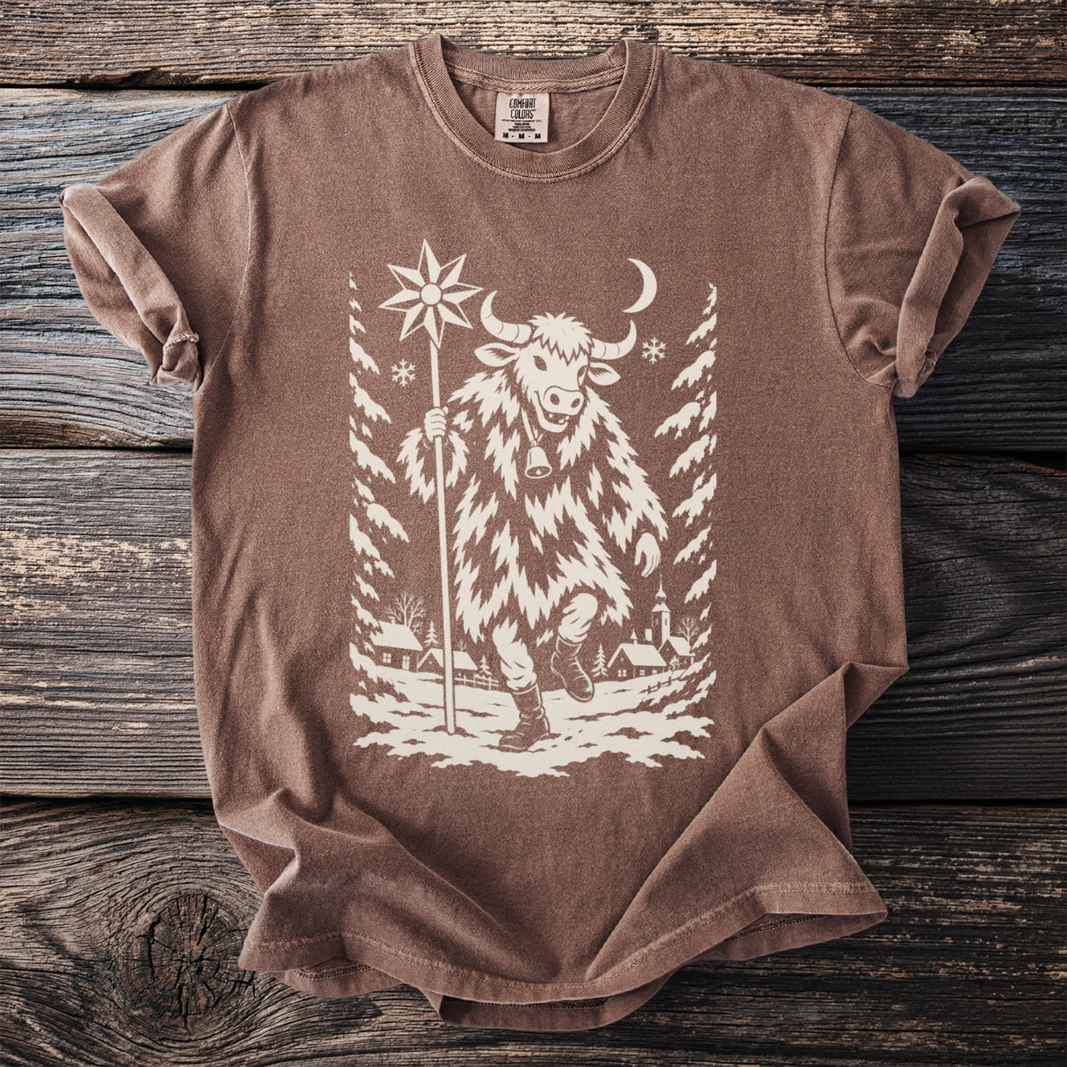 Espresso brown t-shirt with a cream folk-cut print: a bull-masked figure in a shaggy coat walks through a snowy village carrying a star on a pole.