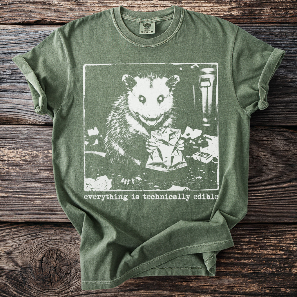 Green t-shirt with white graphic of a possum holding a crumpled bag and text "everything is technically edible."