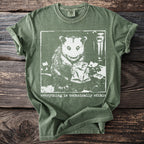 Green t-shirt with white graphic of a possum holding a crumpled bag and text "everything is technically edible."