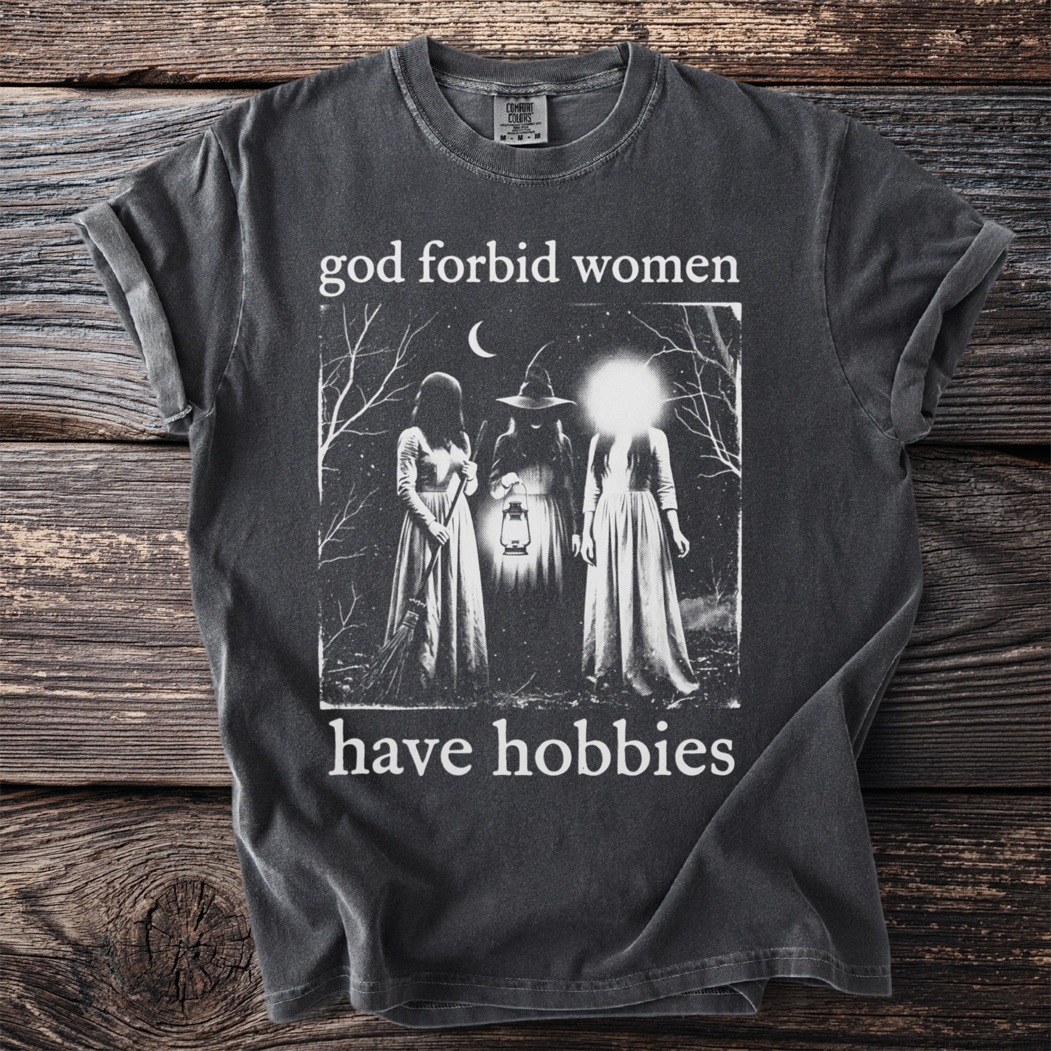 Dark t-shirt with "god forbid women have hobbies" and a night scene graphic.