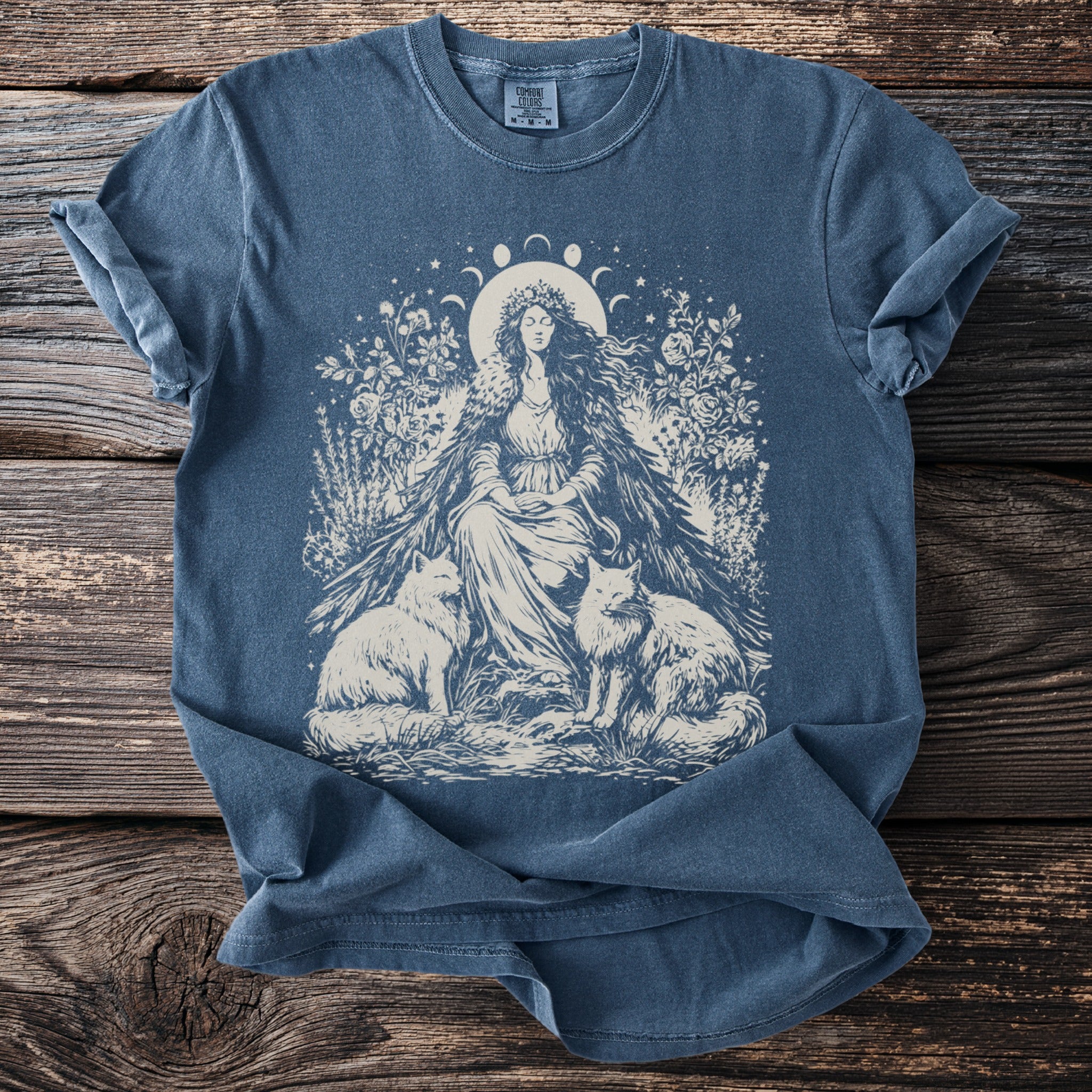 Blue t-shirt with illustration of crowned woman sitting between two foxes and floral elements.