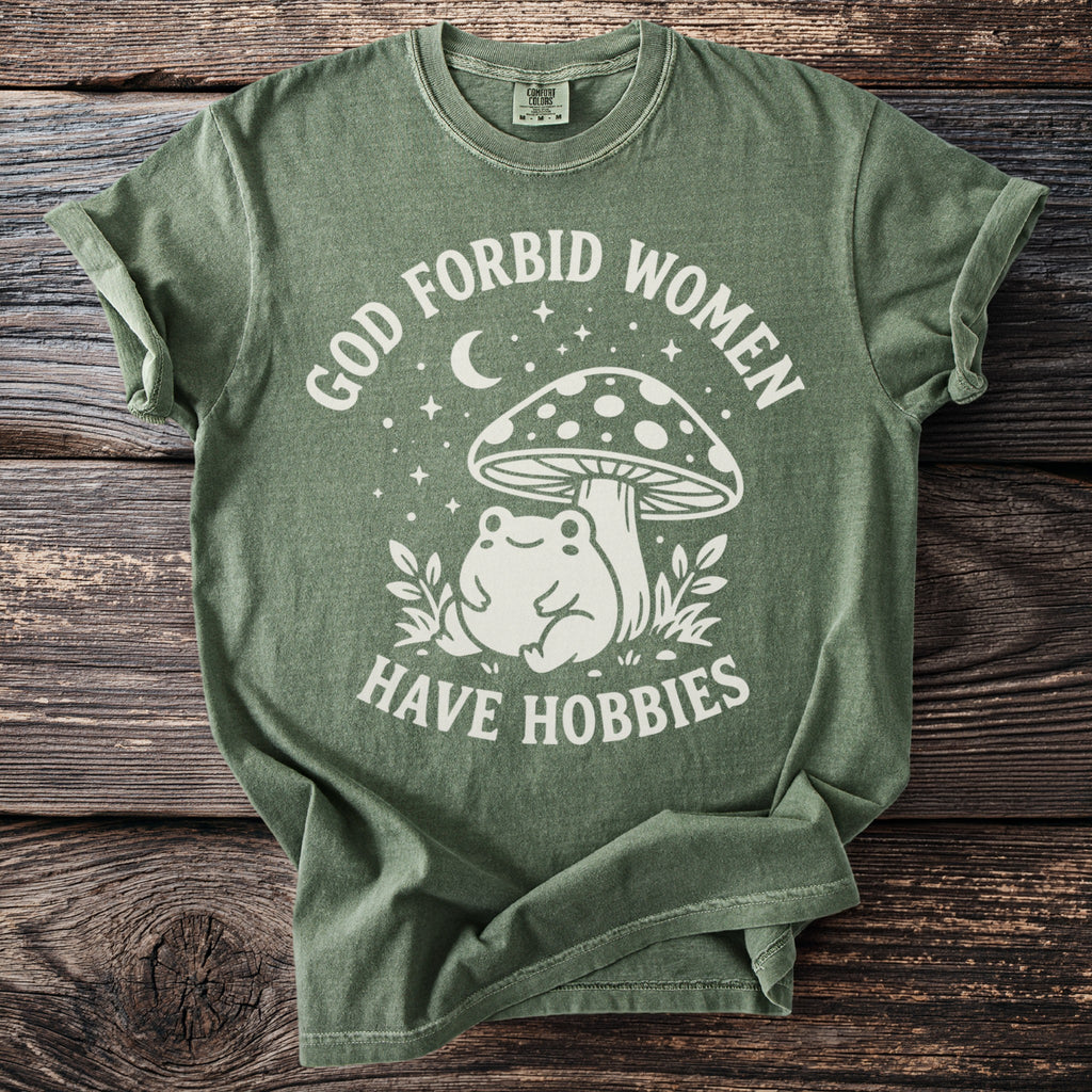 Green t-shirt with white text "God forbid women have hobbies" and a frog under a mushroom.