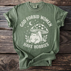Green t-shirt with white text "God forbid women have hobbies" and a frog under a mushroom.