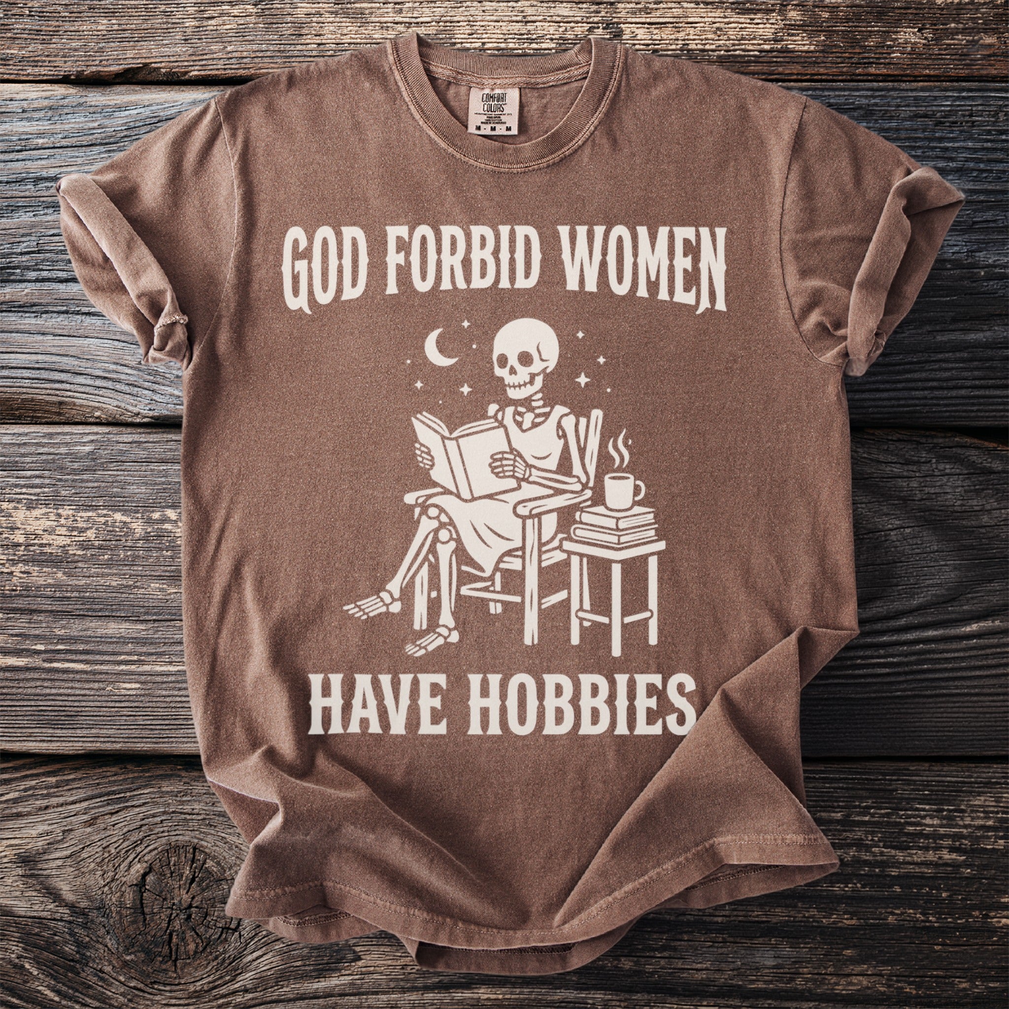 Brown t-shirt with skeleton reading and text "God forbid women have hobbies".