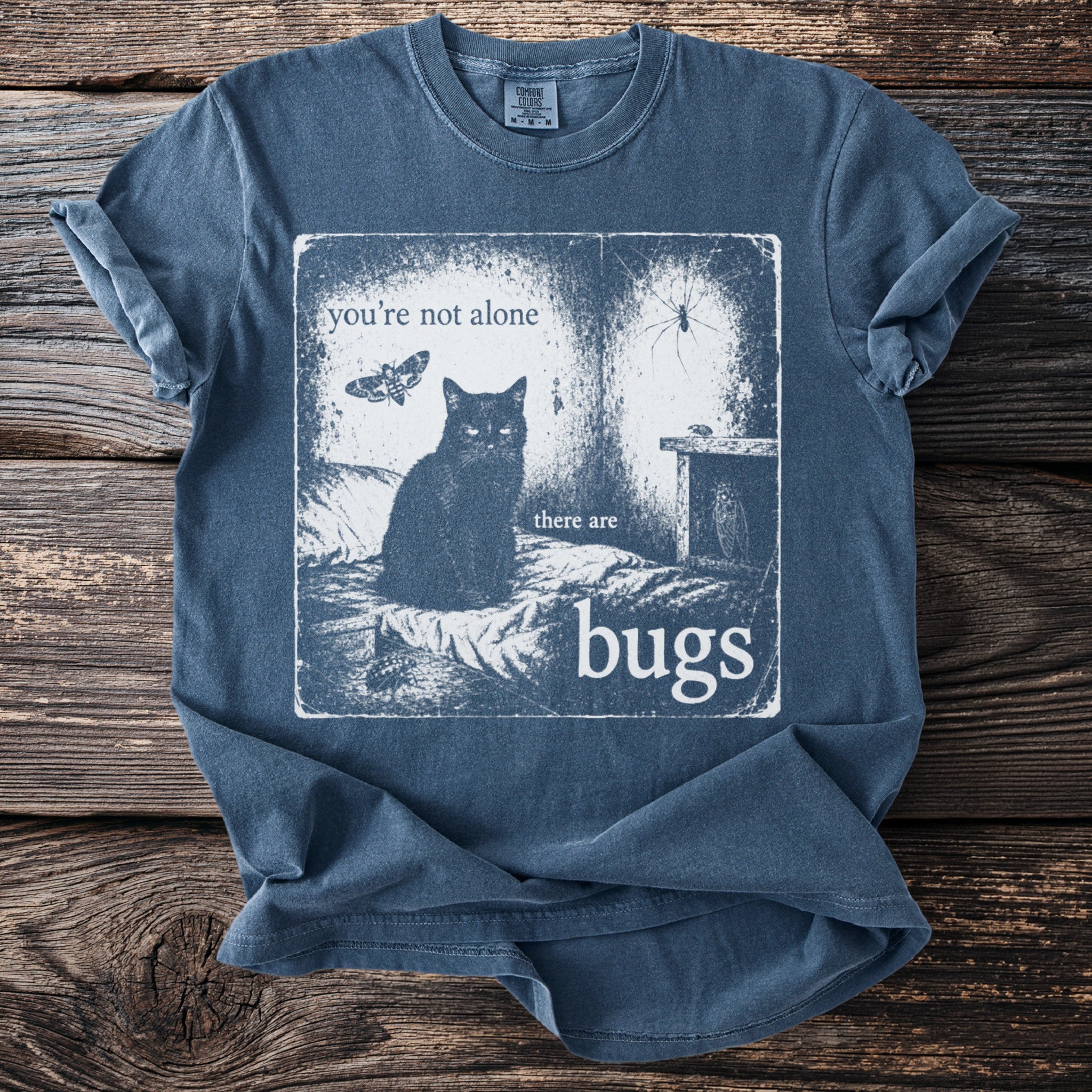 Black cat with moths, spiders, and bugs illustration on a blue shirt reads "you're not alone there are bugs".