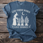 Blue t-shirt with white graphic of witch watering plants and text "God forbid women have hobbies."
