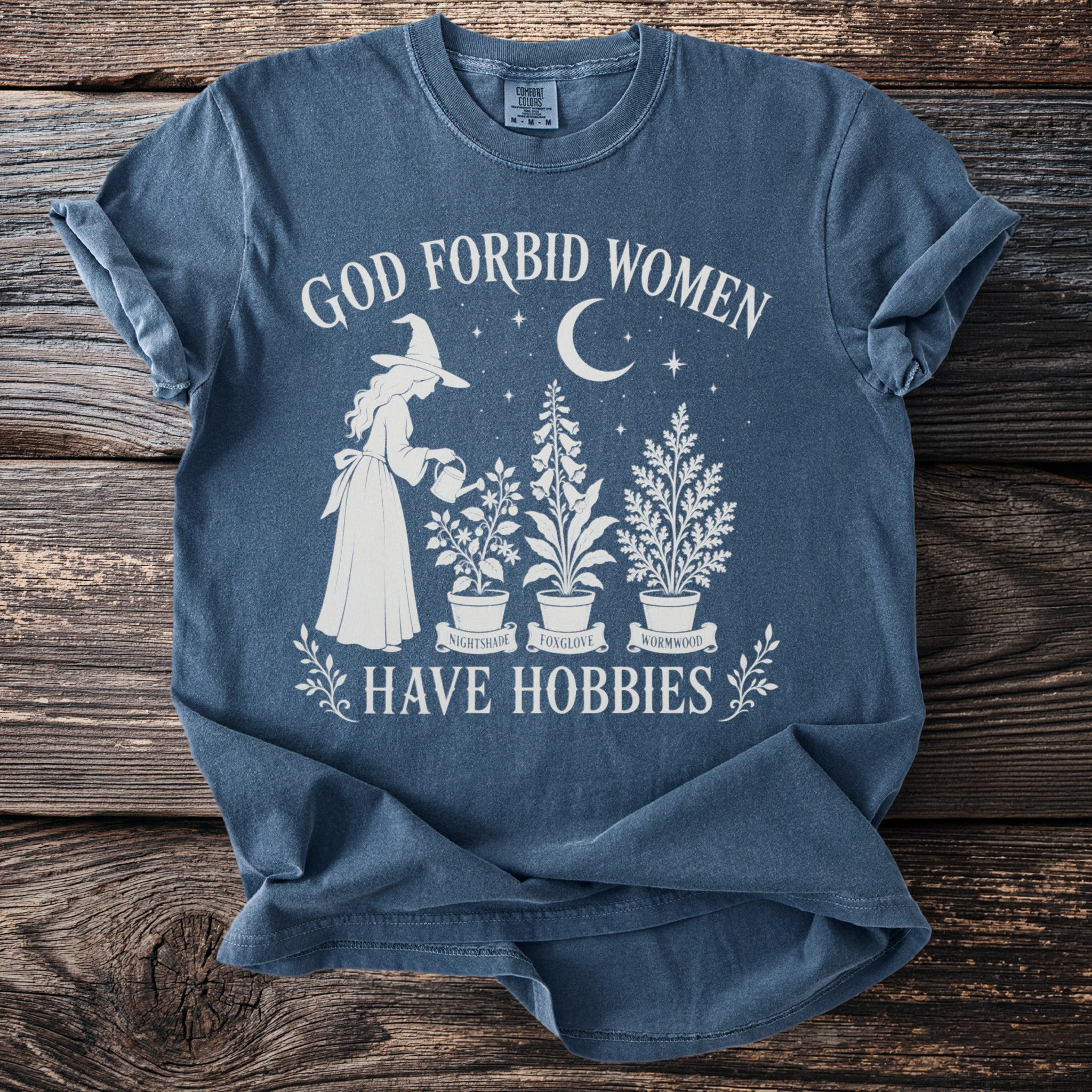 Blue t-shirt with white graphic of witch watering plants and text "God forbid women have hobbies."