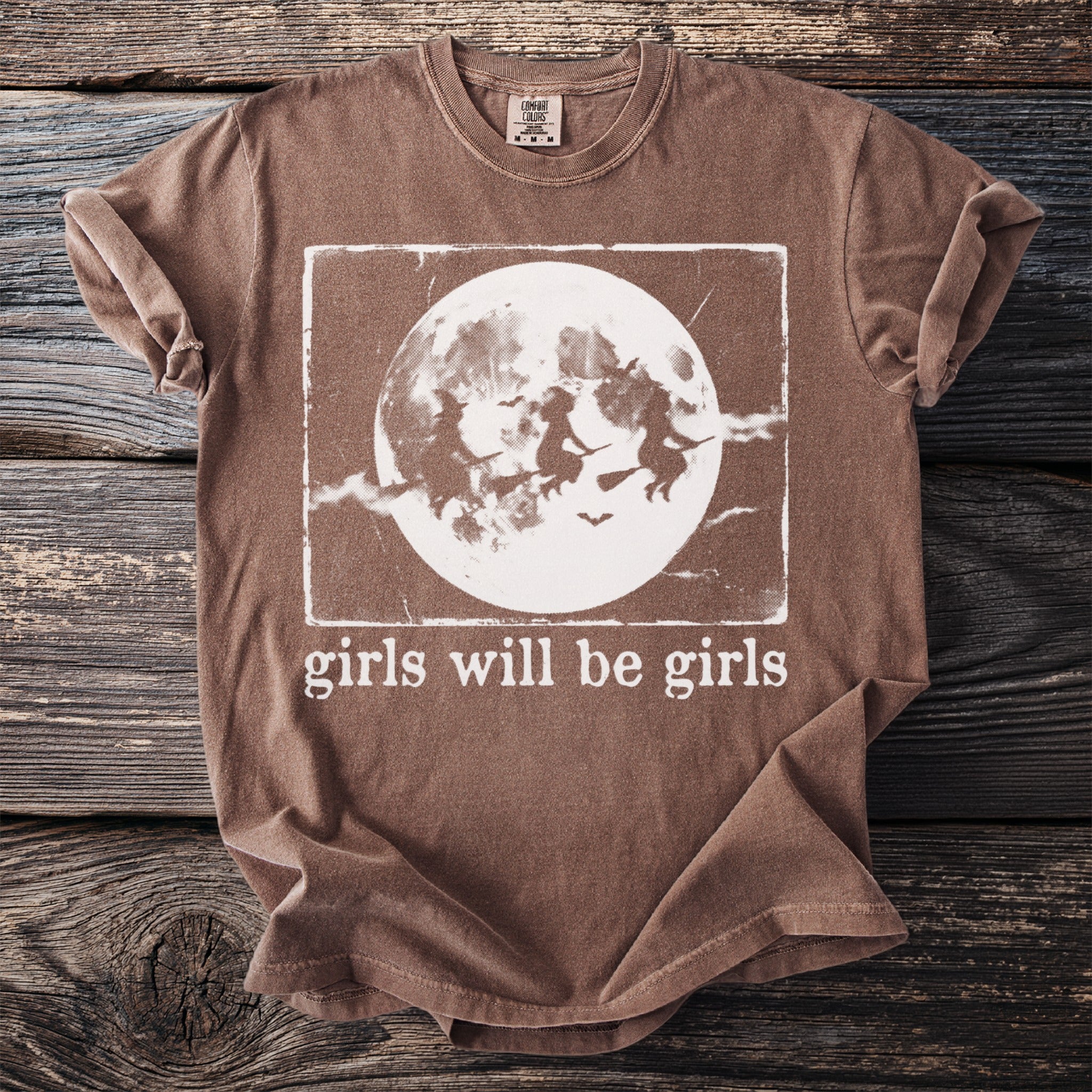 Brown t-shirt with witches flying on broomsticks before a full moon and the text "girls will be girls".
