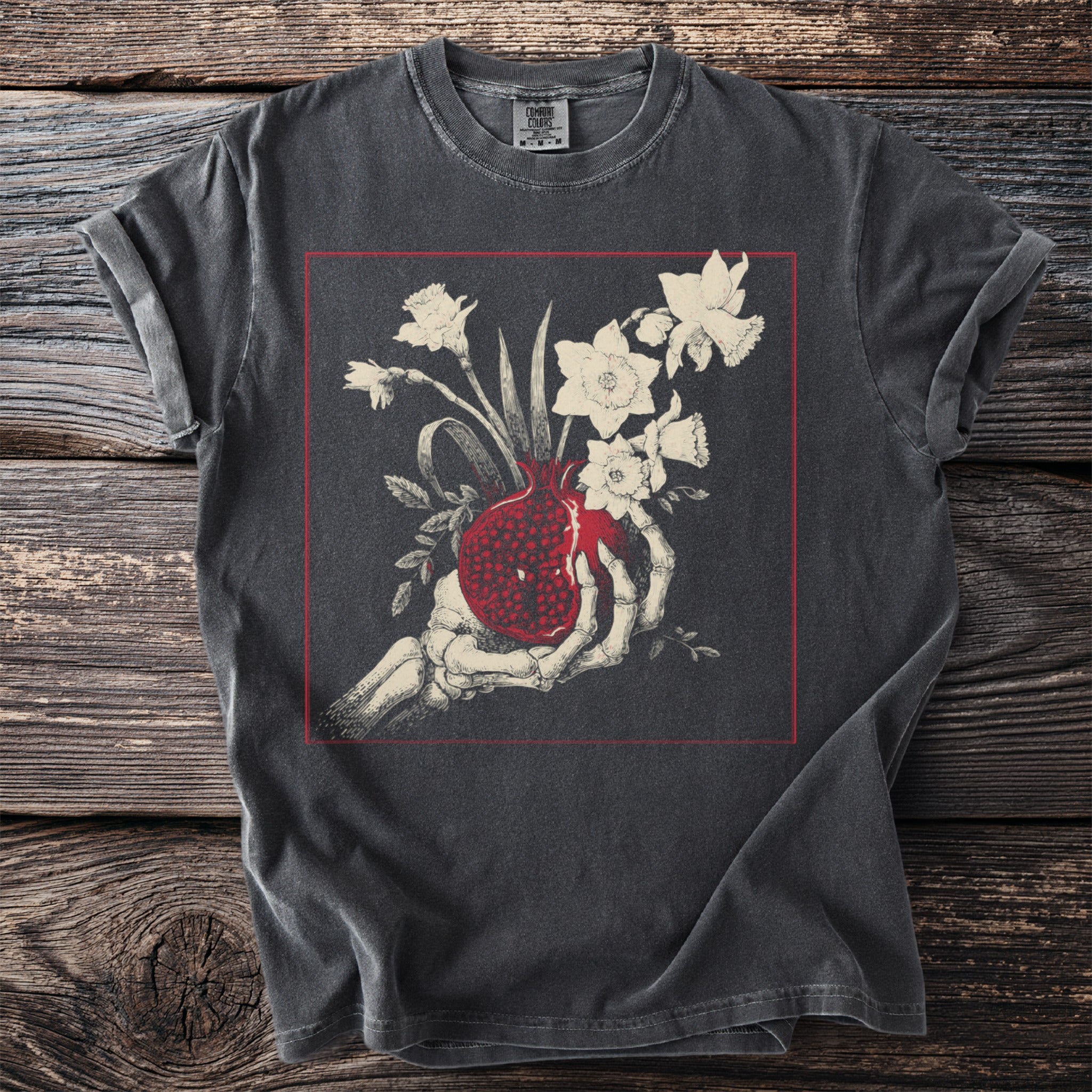 Skeleton hand holding a pomegranate with daffodils within a red square frame.