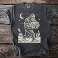 Pepper gray t-shirt with a cream folk-cut print: Bigfoot walks through the snow with an armful of split logs, a cabin chimney smoking behind him.