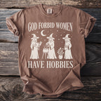 Brown t-shirt with white graphic of three witches and text "GOD FORBID WOMEN HAVE HOBBIES".