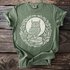 Green t-shirt with white graphic of owl on books within an ornate floral wreath.