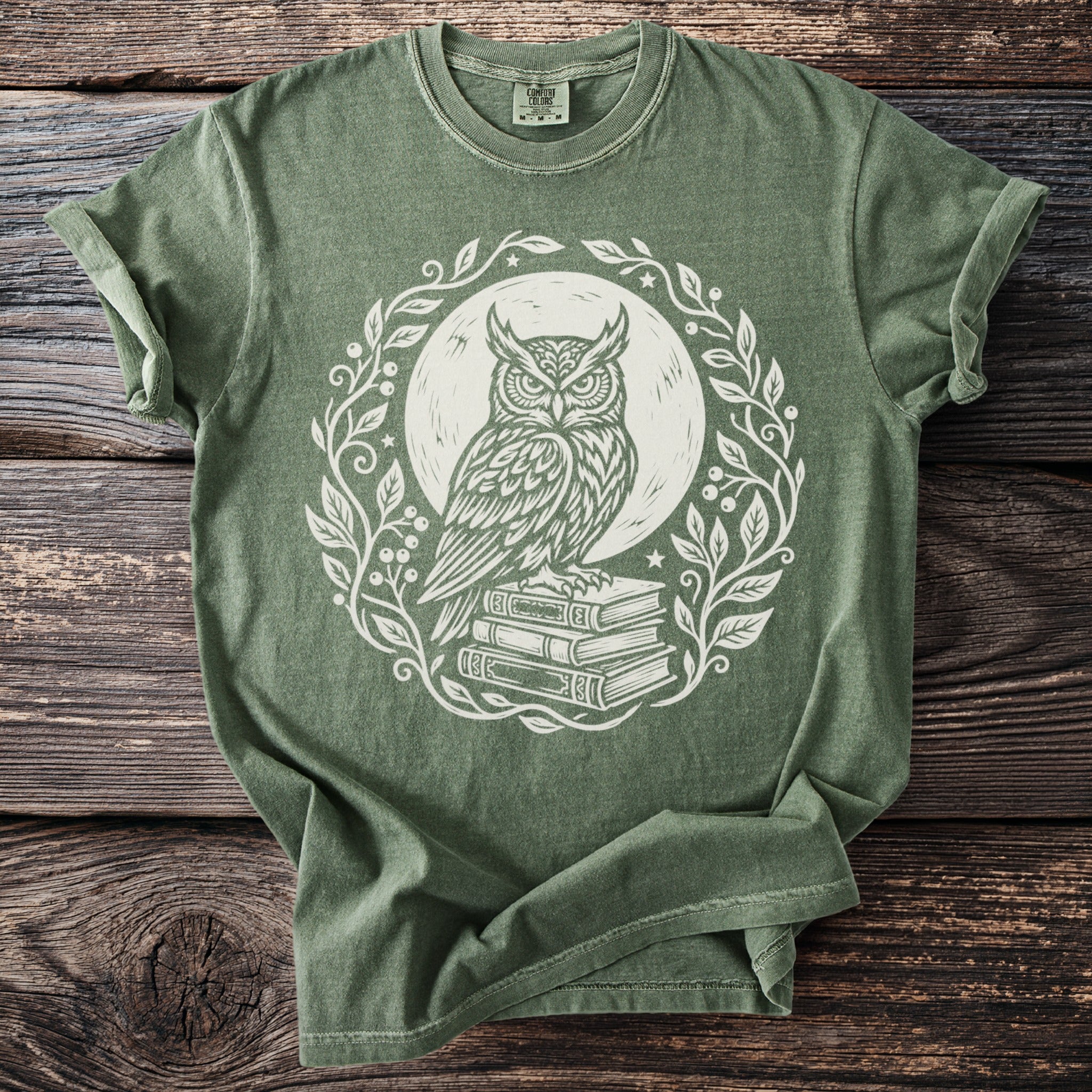 Green t-shirt with white graphic of owl on books within an ornate floral wreath.
