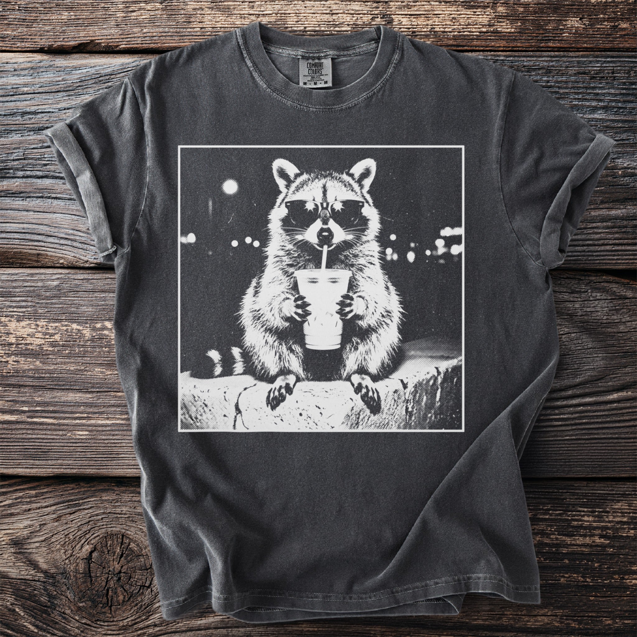 Graphic t-shirt with a black and white raccoon wearing sunglasses and drinking from a cup.