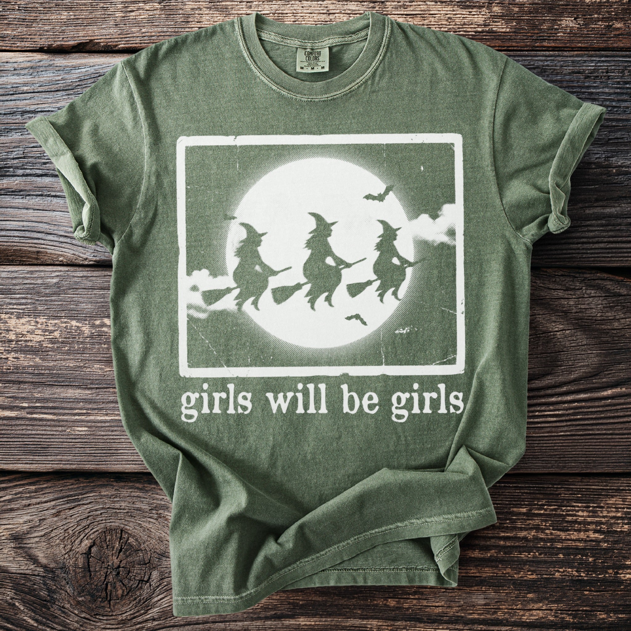 A green t-shirt with three witches riding broomsticks against a moon, and text that reads "girls will be girls".