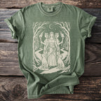 Green t-shirt with a minimalist design of three goddesses in a forest with torches and dogs.