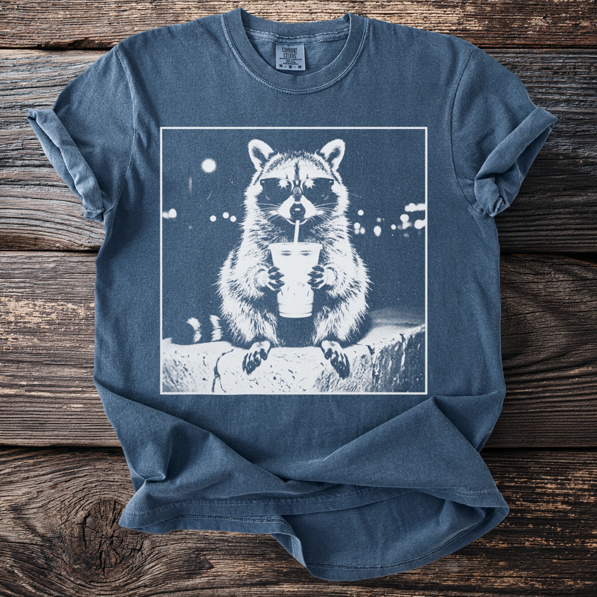 T-shirt with graphic of raccoon wearing sunglasses drinking from a cup.