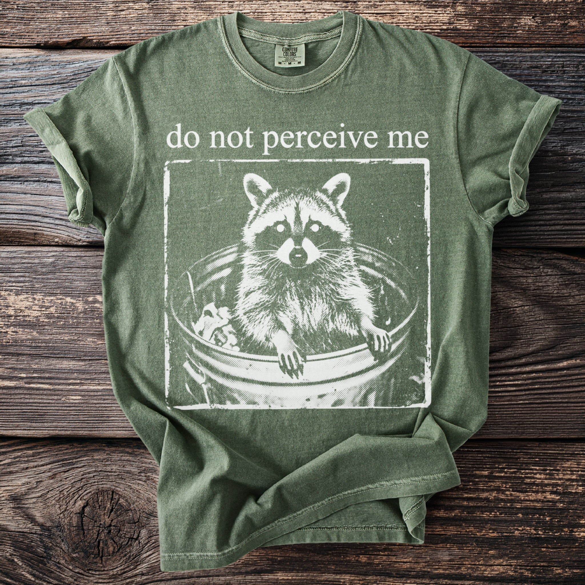 Green t-shirt with white graphic of raccoon in trash can and text "do not perceive me".