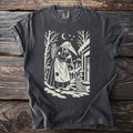 Pepper gray t-shirt with a cream folk-cut print: a beaked Perchta in an apron sweeps a snowy doorstep, scissors in the basket on her back.