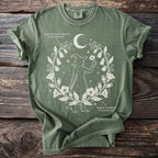 Green t-shirt with witch silhouette, crescent moon, and botanical illustrations of Deadly Nightshade and Black Henbane.
