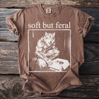 Brown t-shirt with "soft but feral" text above a werewolf looking at kittens in a box.
