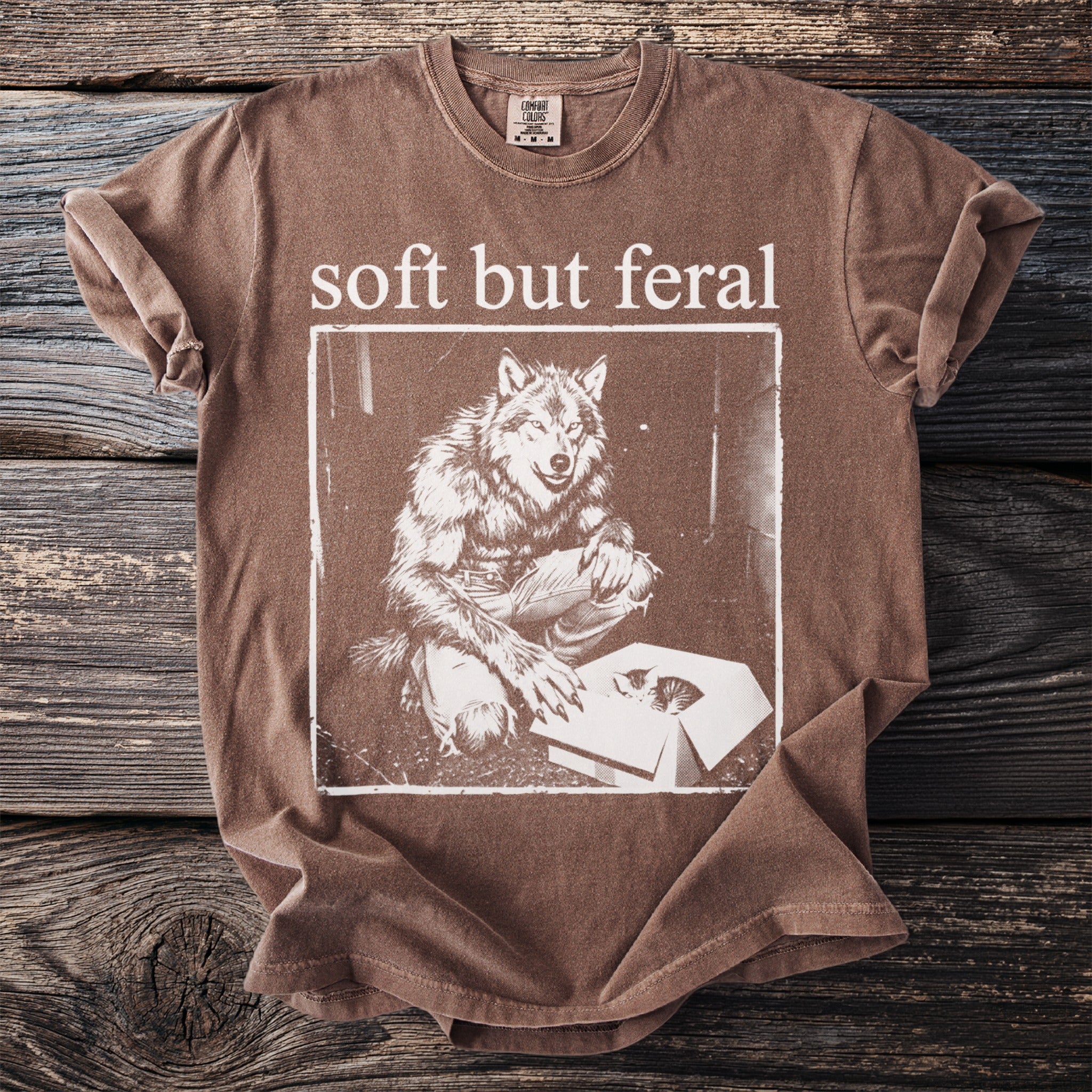 Brown t-shirt with "soft but feral" text above a werewolf looking at kittens in a box.