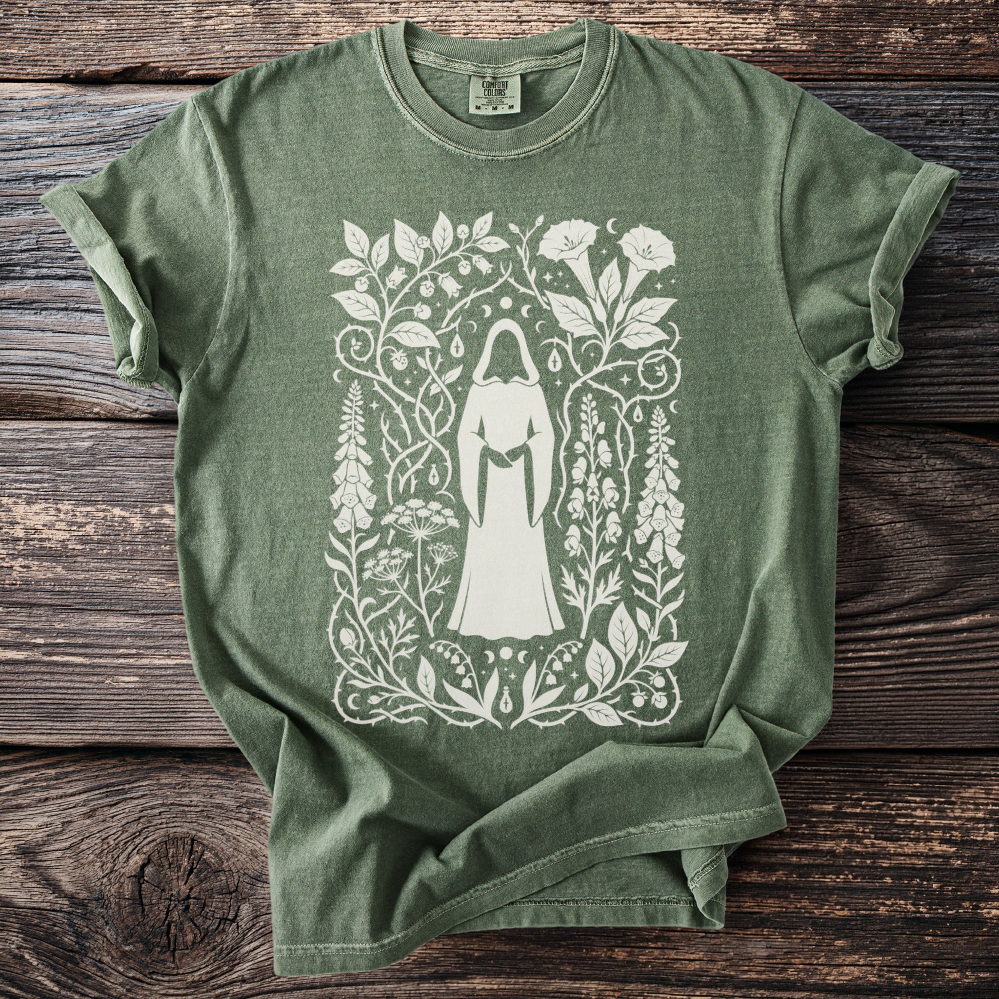 Green t-shirt with a white illustration of a cloaked figure surrounded by botanical elements.