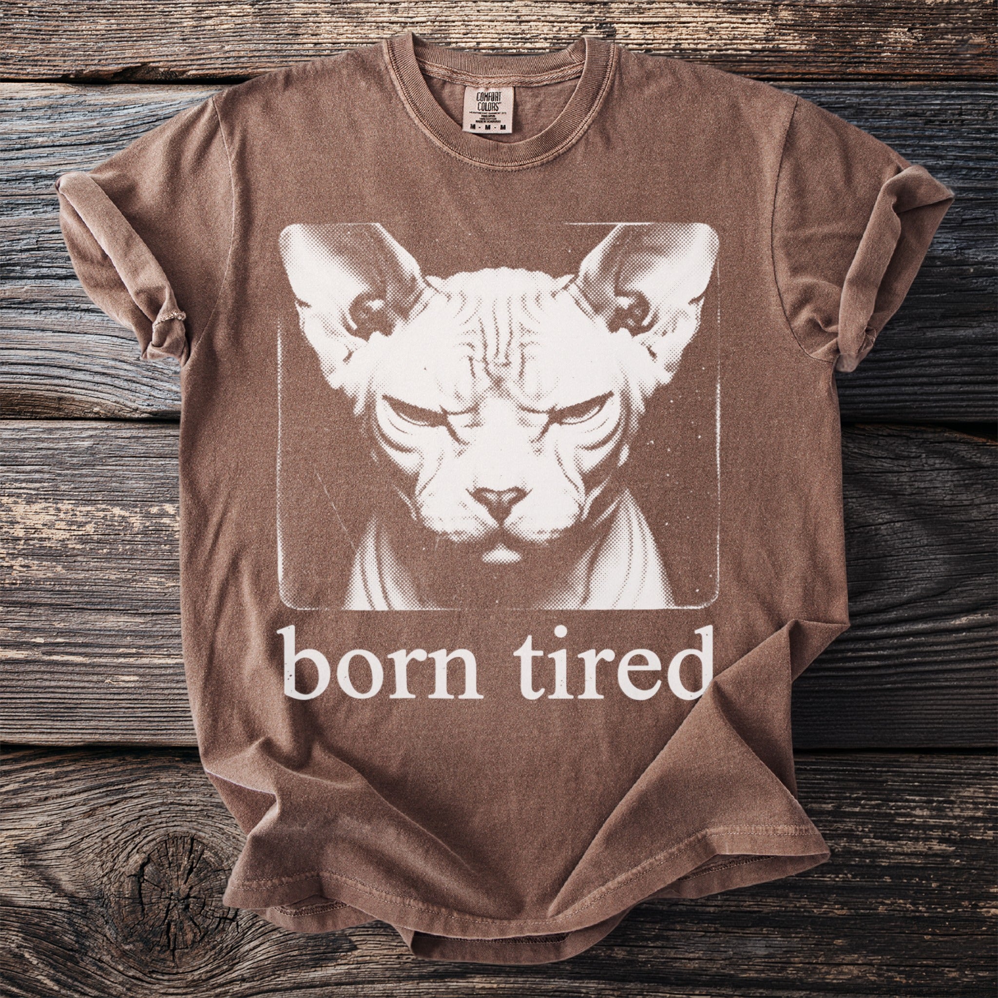 Brown t-shirt with a graphic of a grumpy hairless cat and the text "born tired".