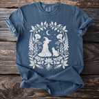 Blue t-shirt with a white graphic of a witch, cat, and celestial elements within floral border.