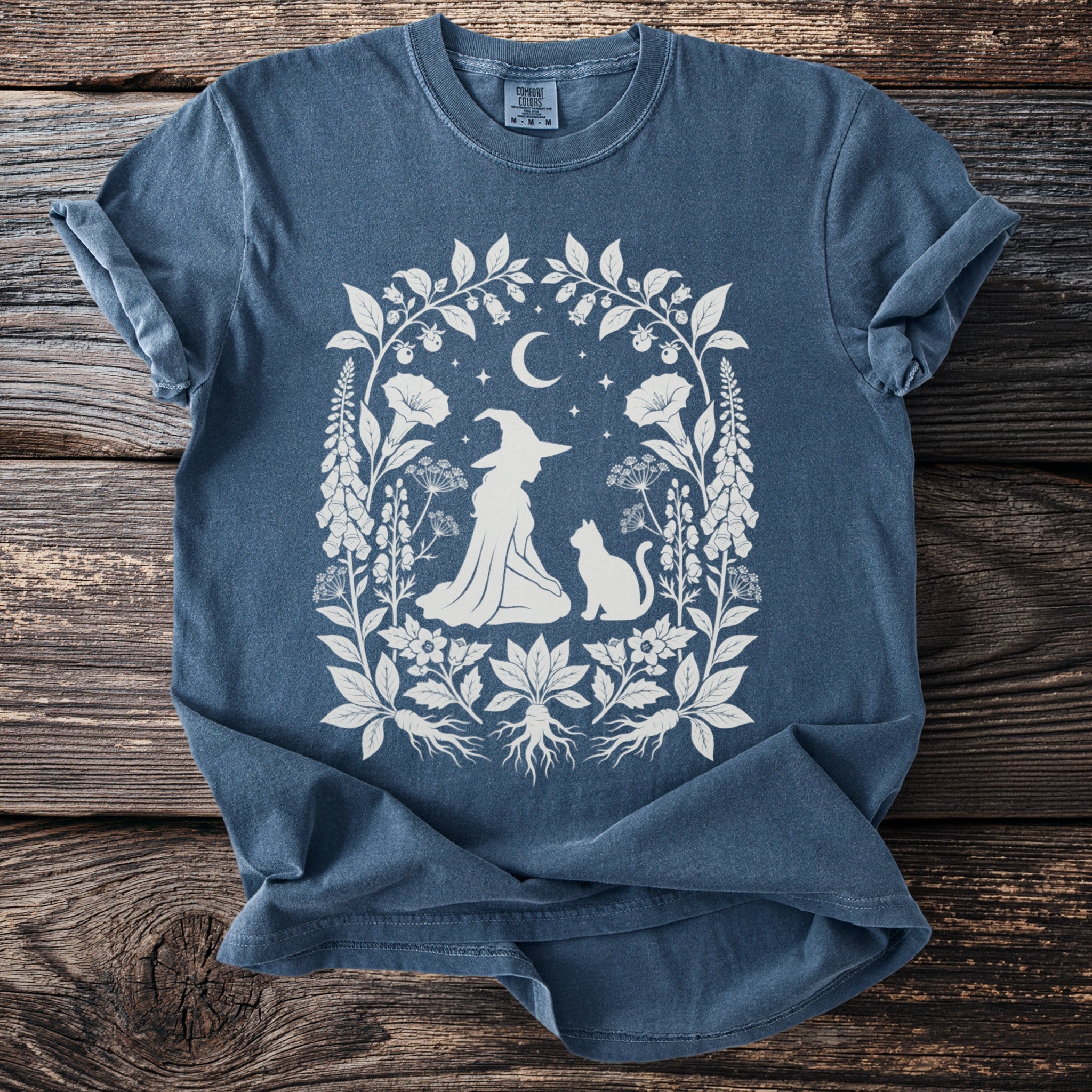 Blue t-shirt with a white graphic of a witch, cat, and celestial elements within floral border.