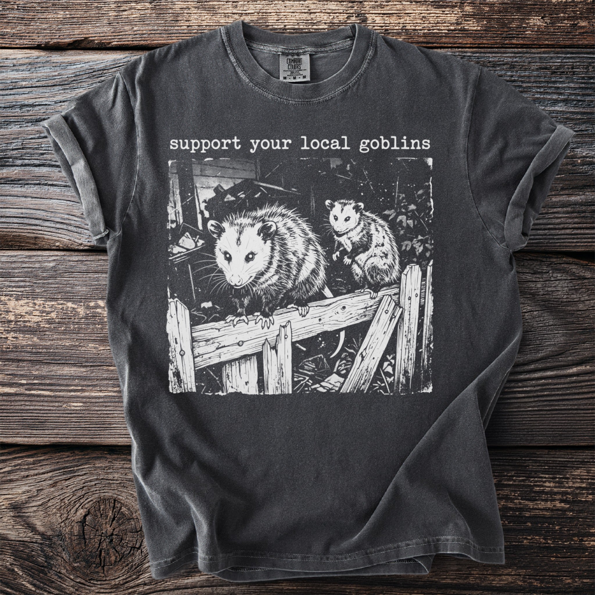 Support your local goblins graphic on a dark gray t-shirt featuring two opossums on a wooden fence.