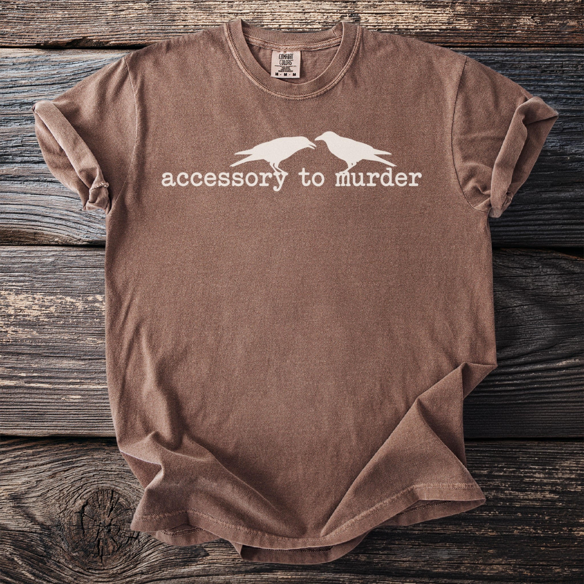Brown t-shirt with white graphic of two birds and text "accessory to murder".