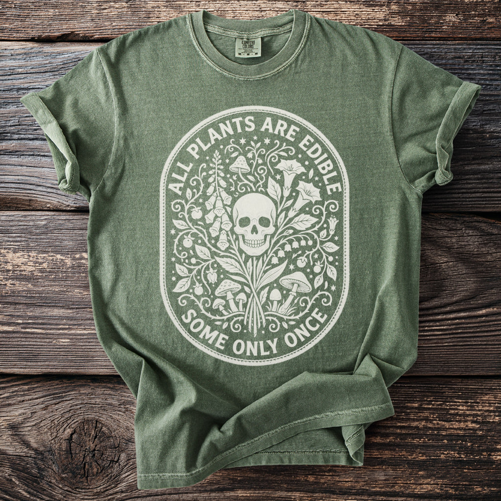 Green t-shirt with white oval graphic featuring a skull, plants, mushrooms, and text "ALL PLANTS ARE EDIBLE SOME ONLY ONCE".